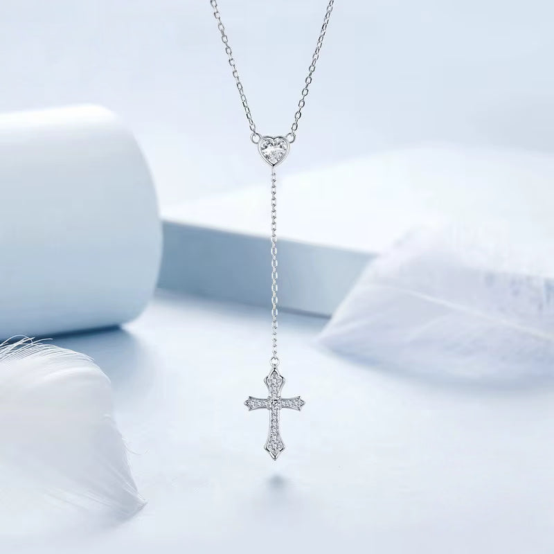 Y lariat silver cross necklace with a CZ heart connector, hanging against a pale blue backdrop
