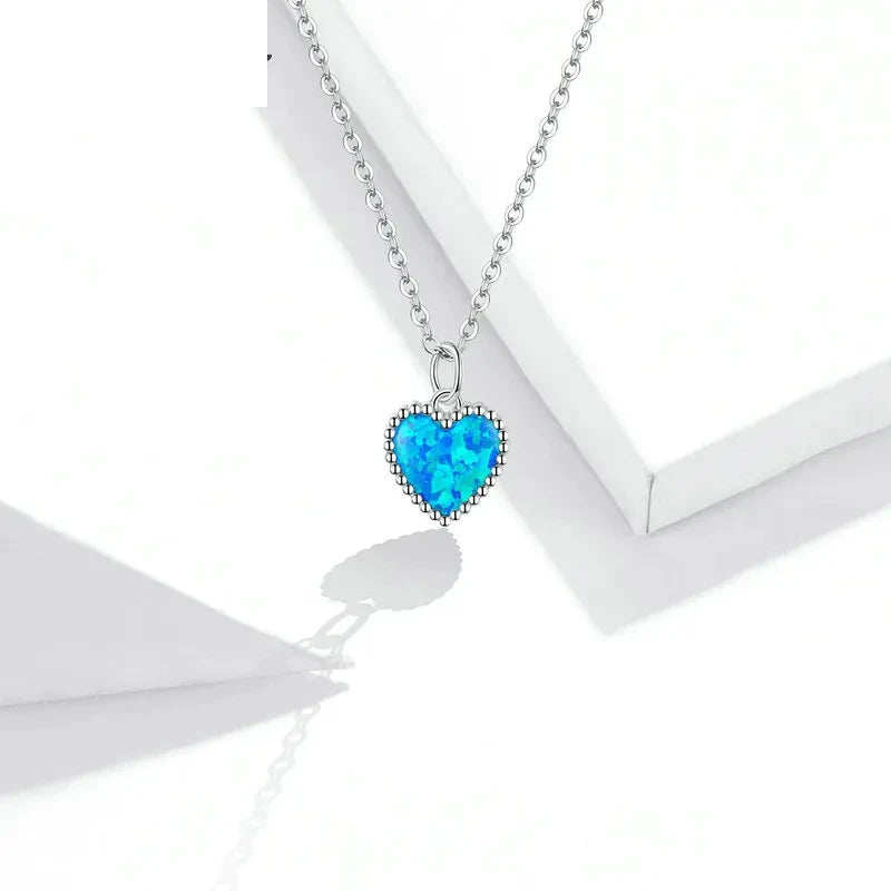 Blue opal-look heart pendant with a silver beaded border displayed against white geometric props