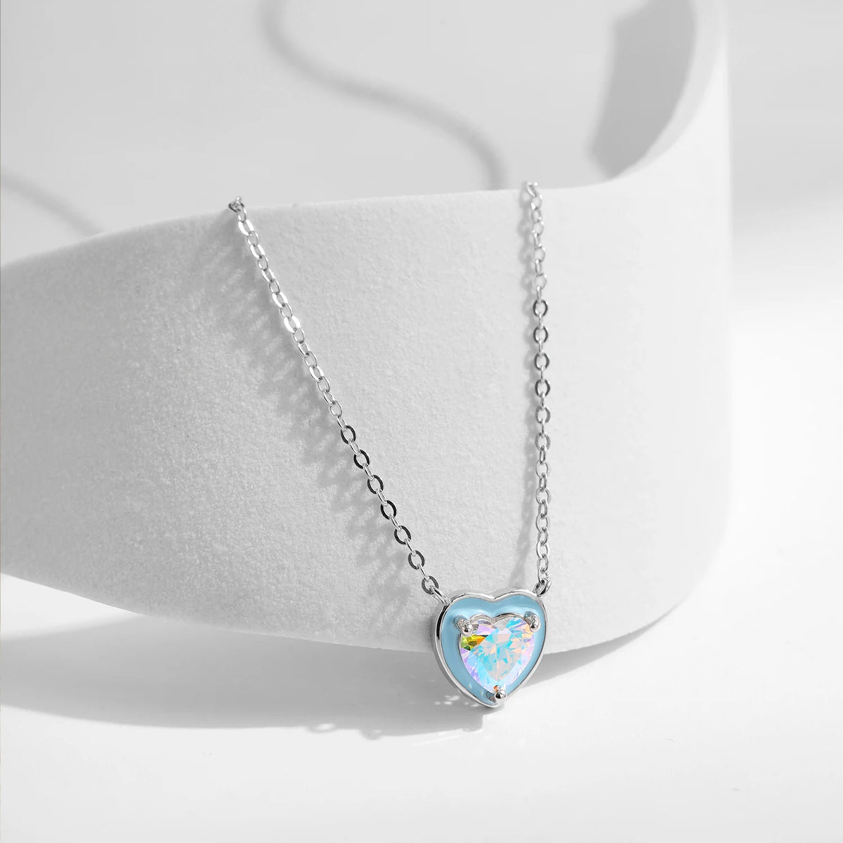 Pale blue enamel heart pendant with a faceted multicolored CZ stone displayed on a white curved surface