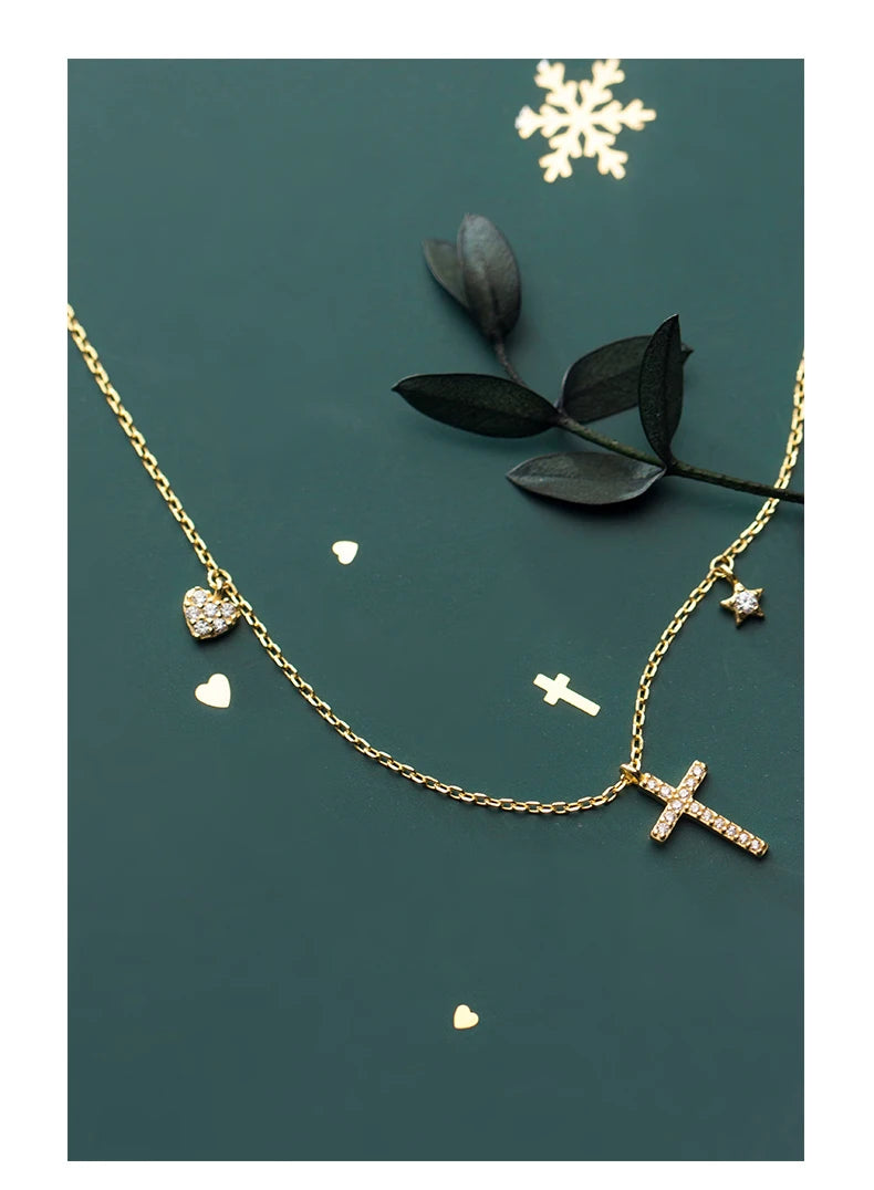 Gold-tone necklace with stone-set heart, tiny star, and cross beside dark leaves