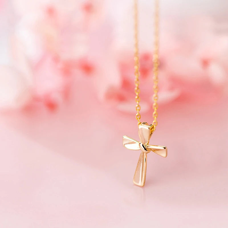 Gold-tone polished Latin cross necklace with gently flared arms suspended against a soft pink backdrop