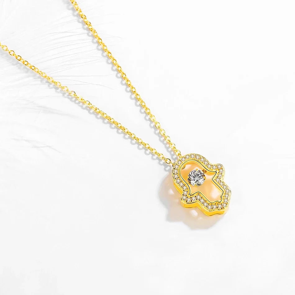 Gold-tone open hamsa hand necklace laid on a white textured surface with clear stone outline