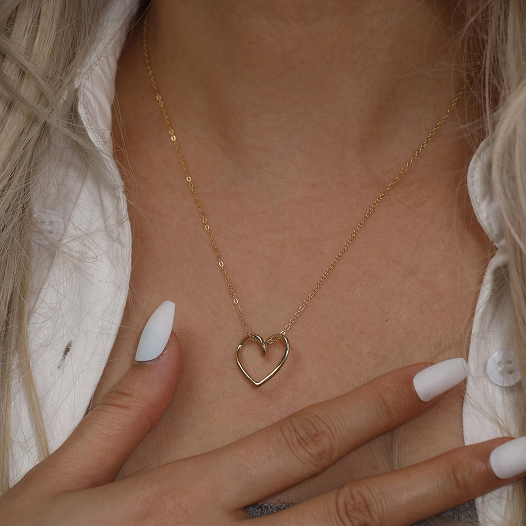 Gold-tone heart outline necklace worn at the base of the throat with a hand in view