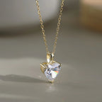 Gold-tone heart necklace hanging over a neutral surface, showing the clear faceted stone and three-point setting