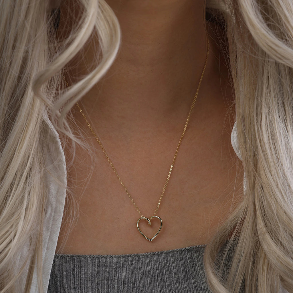 Gold-tone open heart necklace worn on the chest above a gray ribbed top