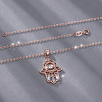Rose gold-colored hamsa necklace laid on gray fabric, showing the oval-link chain, clasp and pink stone evil eye