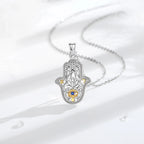 Silver-colored hamsa hand necklace arranged on white fabric with a blue eye and gold-colored accents