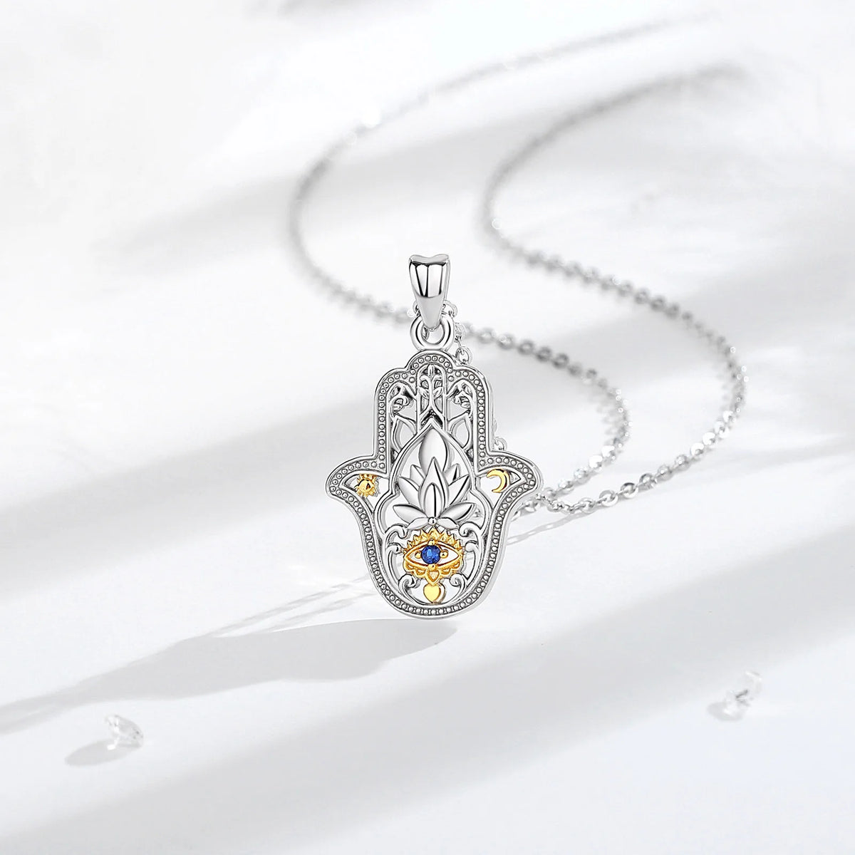 Silver-colored hamsa hand necklace arranged on white fabric with a blue eye and gold-colored accents
