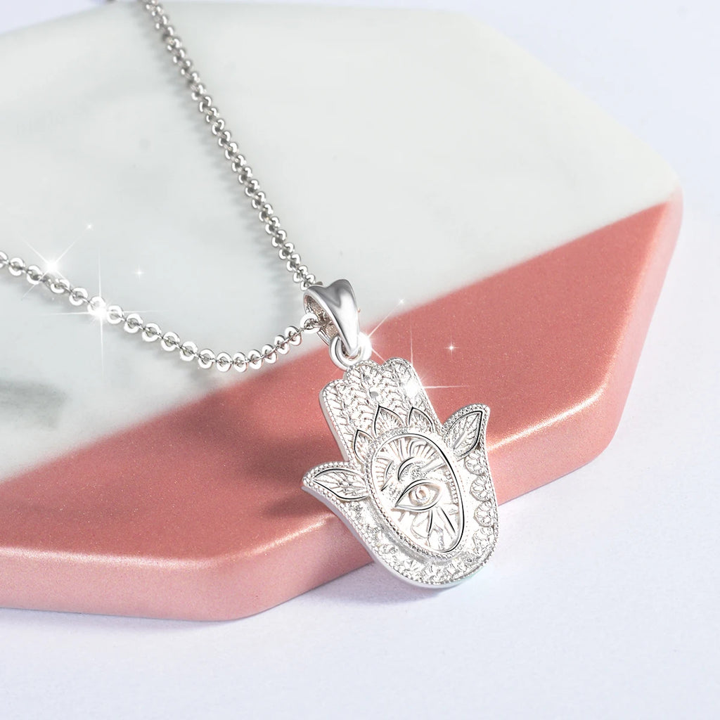 Silver hamsa pendant necklace draped over pink and white display blocks with a raised eye center
