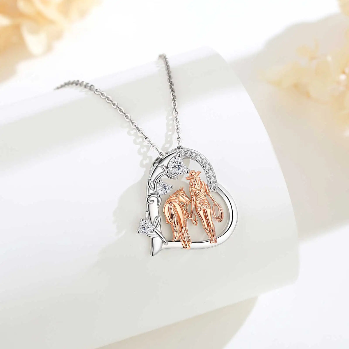 Silver-colored heart necklace with rose-gold-colored cowgirl and horse pendant resting on a white curved display