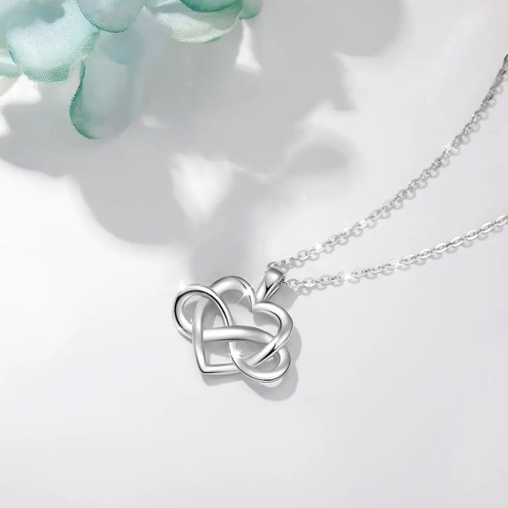 Polished sterling silver heart and infinity necklace arranged on a white surface beside pale petals