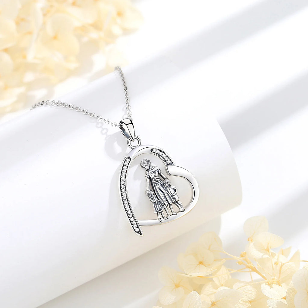 Open heart pendant with mother and two children resting on a white display block beside pale flowers