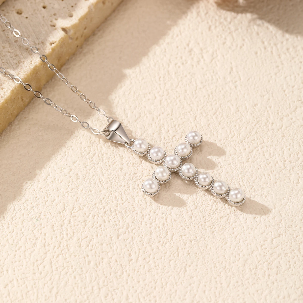 Pearl cross pendant lying on a textured cream surface with its oval-link silver chain