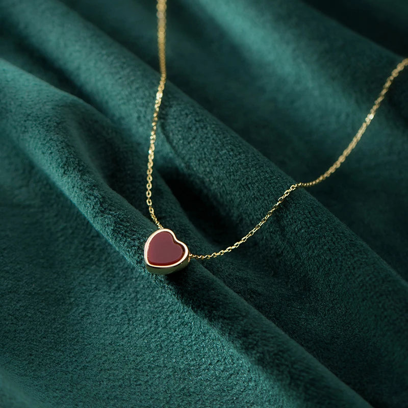 Red heart pendant in a gold-colored rim laid across dark green textured fabric with a fine chain