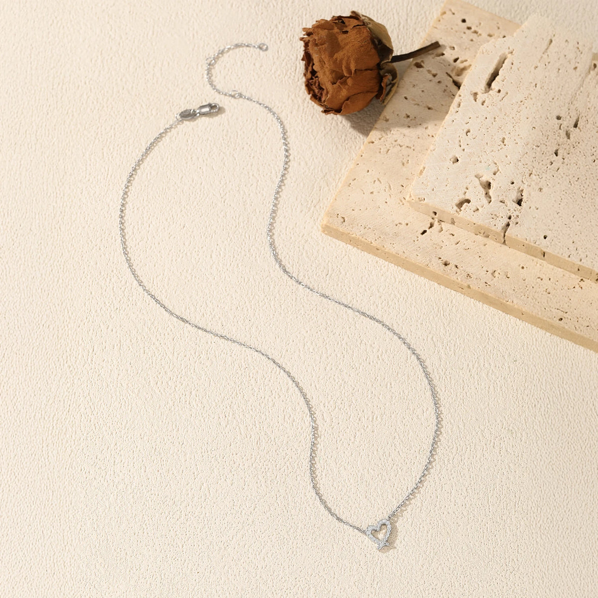 Sterling silver ribbon heart necklace arranged beside a dried flower and pale stone blocks