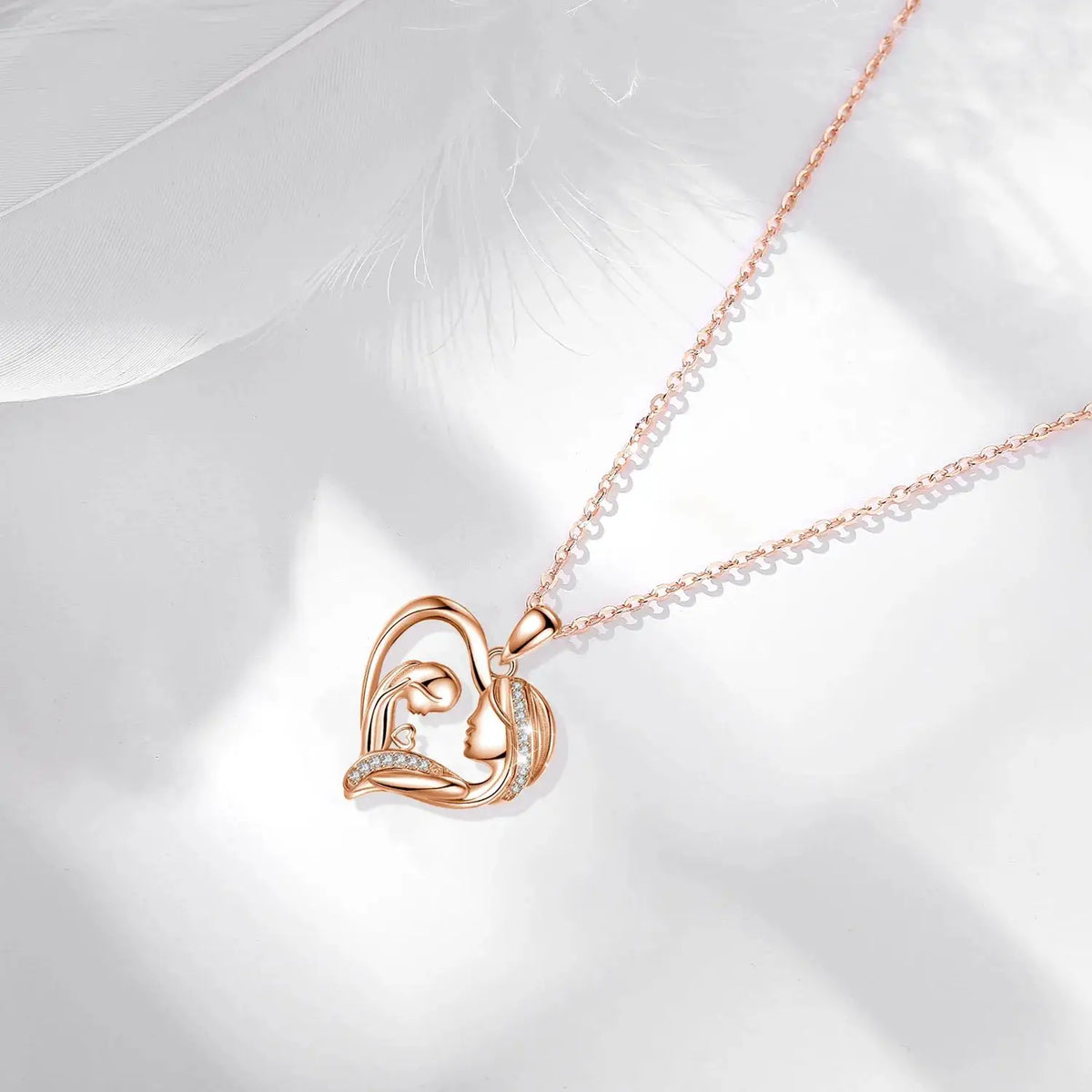Rose gold-tone mother and child heart necklace arranged on a white surface with a feather