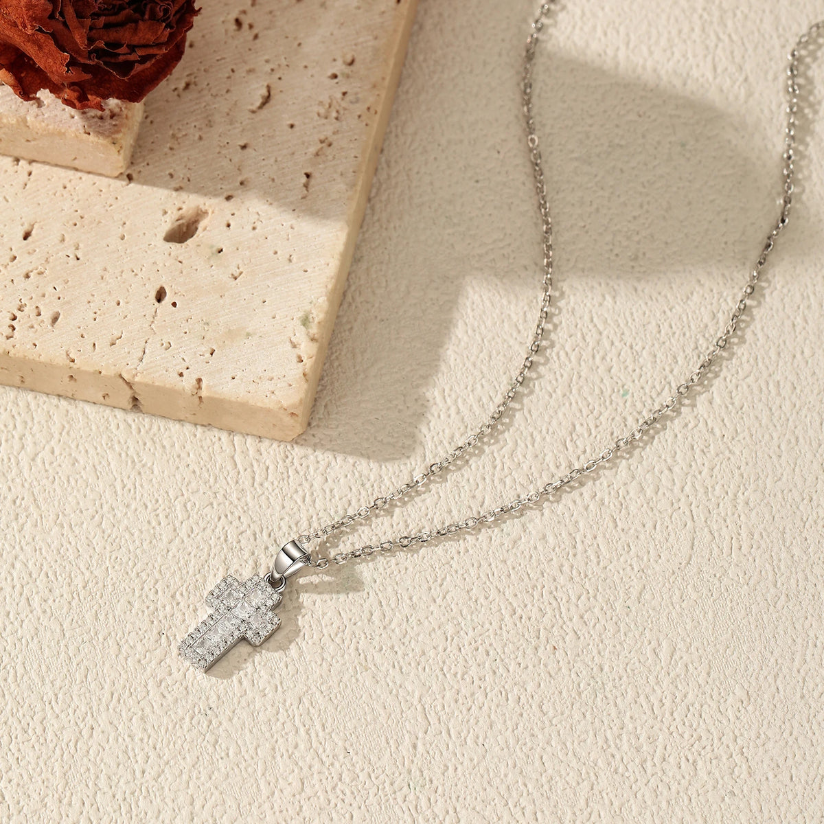 Silver-tone cross necklace arranged on a textured surface, showing the fine oval-link chain