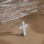 Silver-tone moissanite cross pendant hanging beneath a clear glass prop on a warm surface