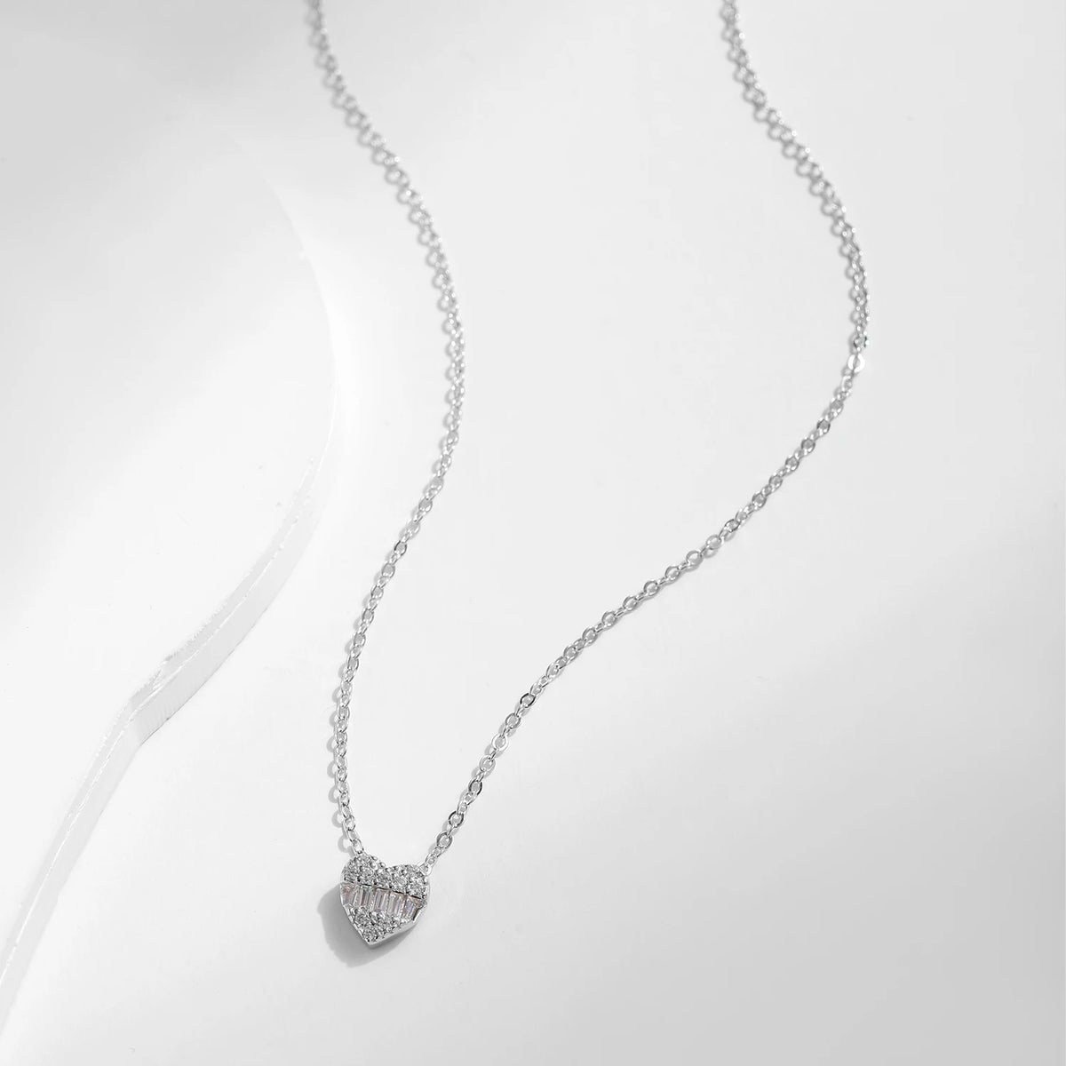 Silver heart necklace displayed on a white curved surface, showing the fine cable chain and stone-set pendant