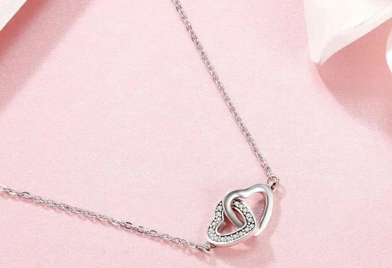Silver-tone interlocking heart pendant with a clear CZ stone row on a pink surface