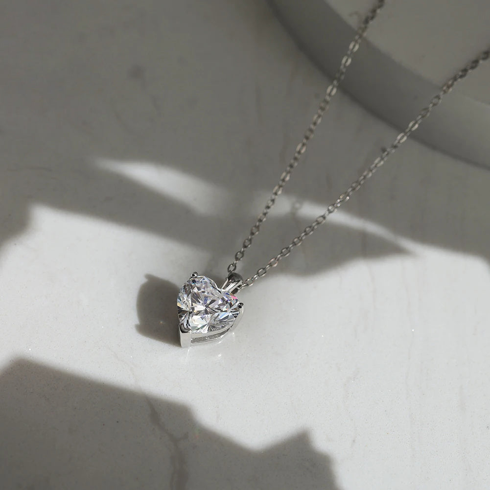Silver-tone heart moissanite pendant lying on a pale marble surface with oval-link chain and side profile