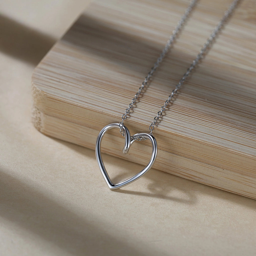 Silver-tone open heart necklace resting beside a light wood block, showing its rounded outline