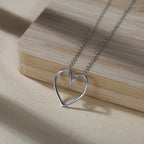 Silver-tone open heart necklace resting beside a light wood block, showing its rounded outline