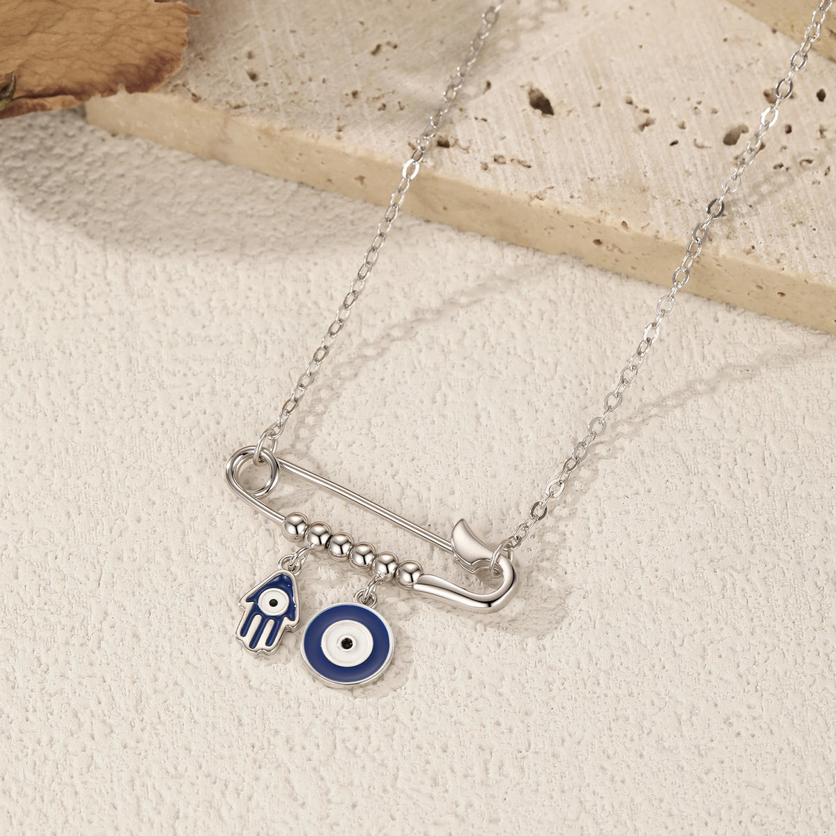 Angled flat lay of the silver safety pin necklace with blue enamel Hamsa and eye charms