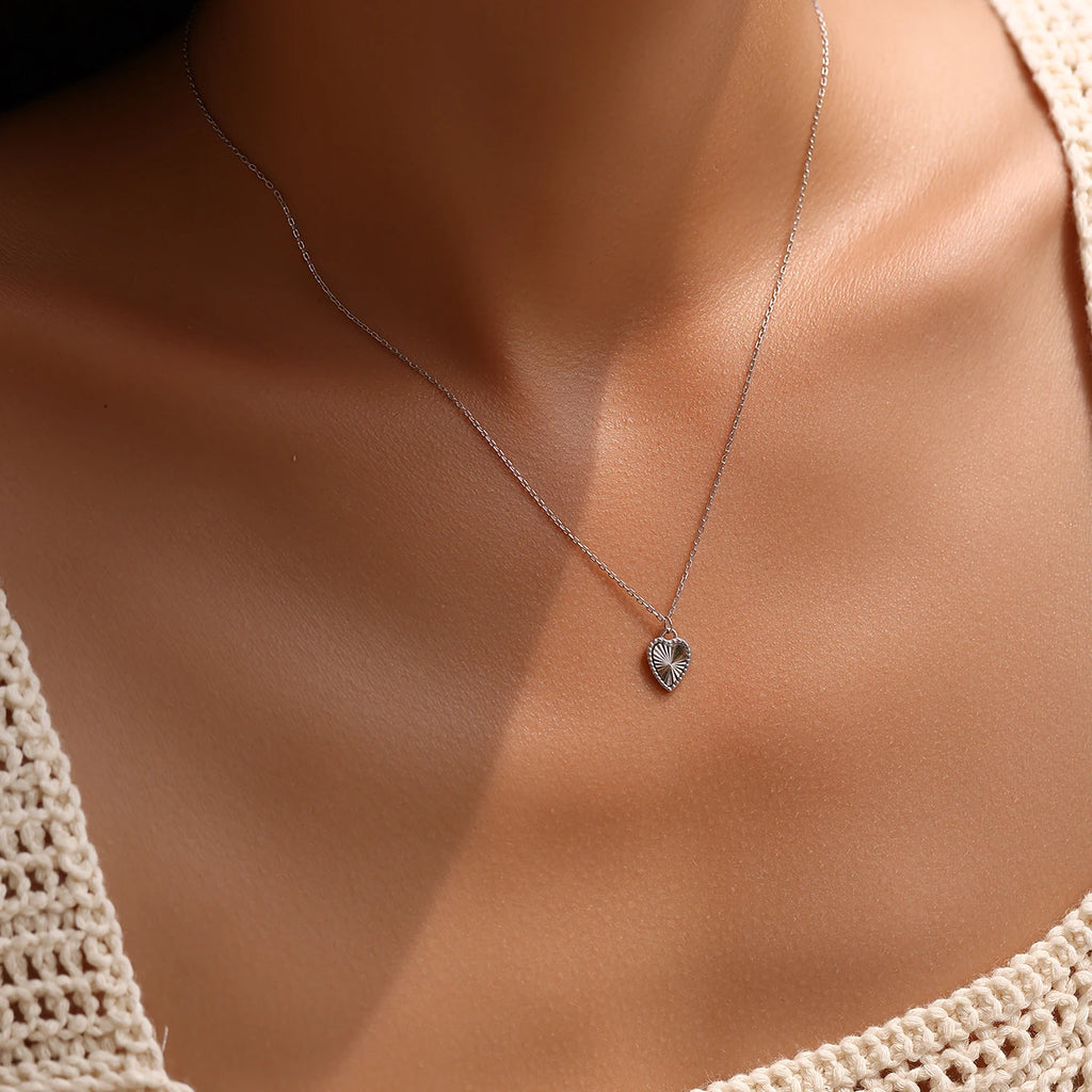 Small sterling silver heart pendant necklace worn at the base of the neck with sunburst rays visible